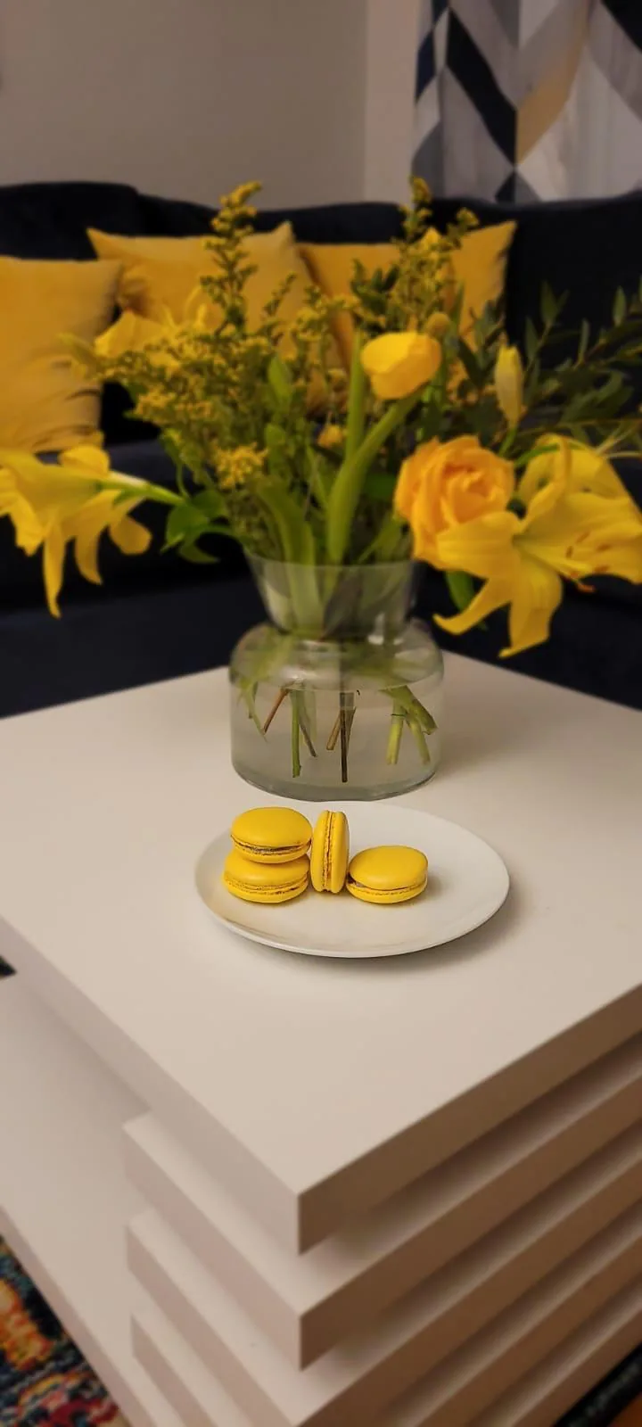 Four yellow lemon macarons on a white plate in front of yellow flowers